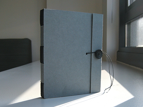 Front view: blue-gray paper cover with black spine stitches and flap fastened with a vintage Japanese black button and linen thread