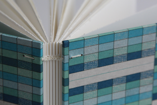 Spine stitch detail of white thread; the cover paper is various blues and turquoises in a rectangular grid pattern